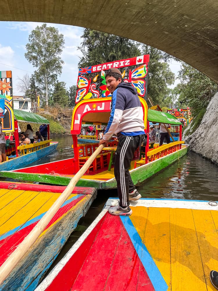 A man in a white shirt and black sweatpants holds a long wooden pole, which he uses to guide a colorful boat. Another colorful boat is in the background