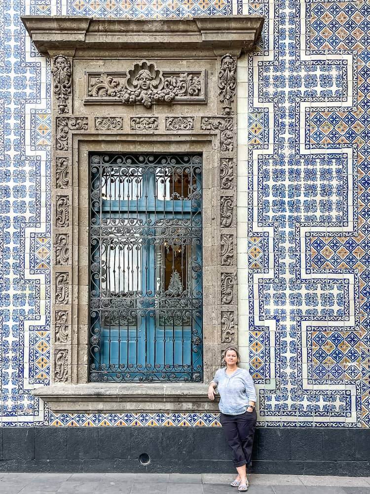 A woman in a light blue shirt and black capri pants stands in front of a large stone window set into a wall covered in elaborate blue and white ceramic tiles.