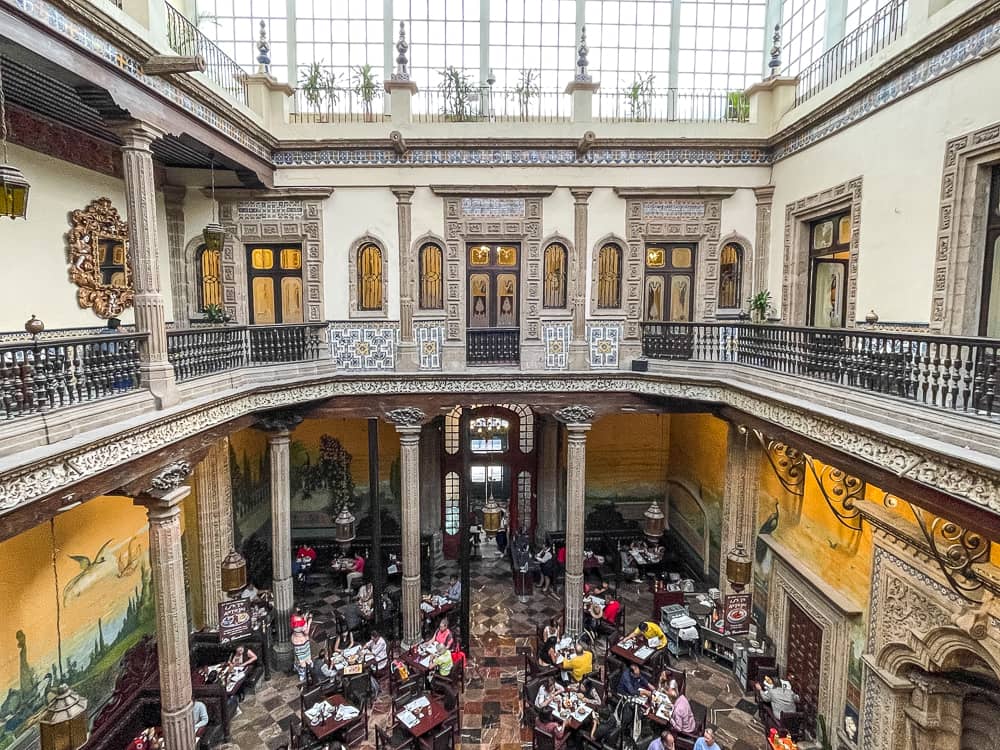 A large two-story restaurant space with tall stone columns and a glass-paned ceiling. The walls on the lower level are bright yellow, and white on the upper level. There are people sitting at tables on the ground floor.