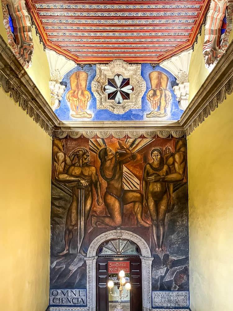 A dark reddish-grey mural depicting scantily clad people sits on a back wall over a stone archway. There is a smaller blue and yellow mural above and yellow walls on the left and right. The ceiling has red beams going from left to right.