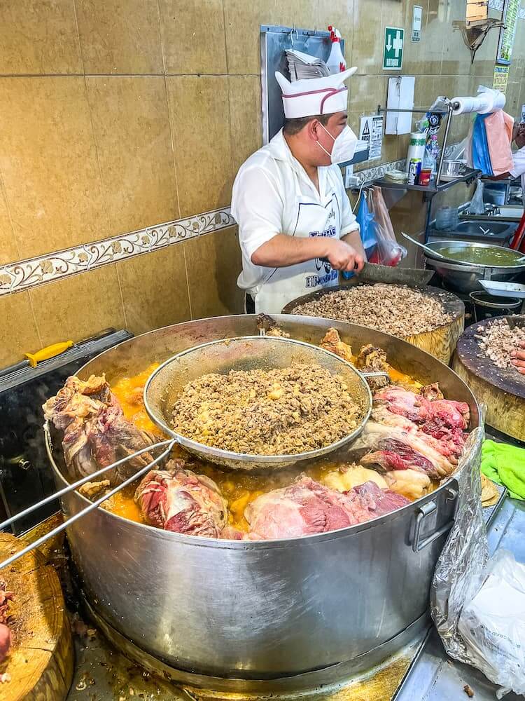 A cook in a white shirt, hat, and apron uses a large knife to chop meat on a wooden block. In the foreground in a large metal pot filled with all kinds of meats.