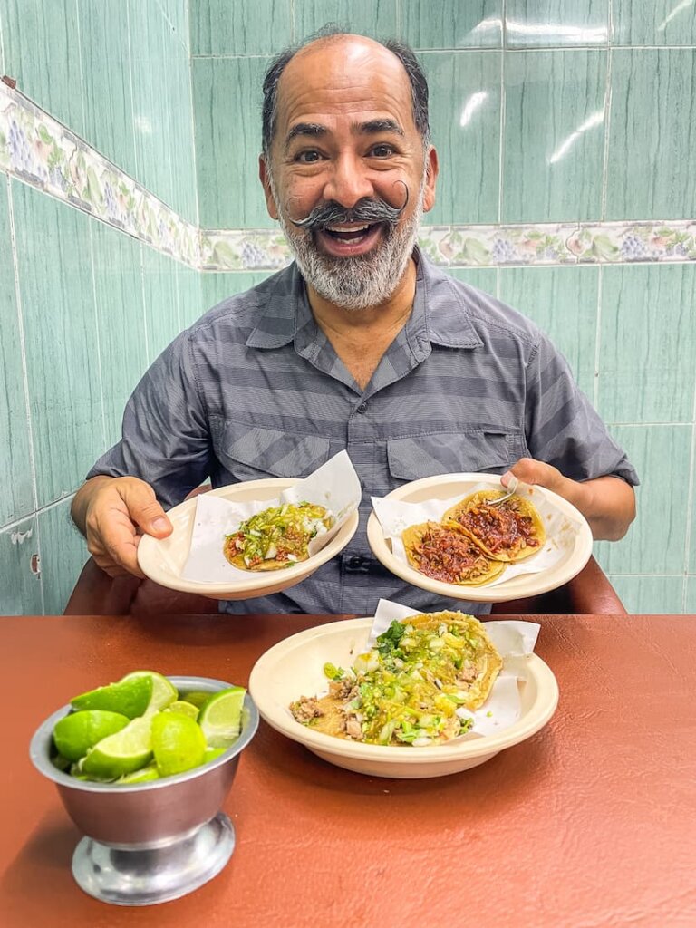 A handsome man with a handlebar mustache holds two plates with tacos. On the orange table in front of the man there is another plate of tacos and a metal bowl filled with sliced imes.