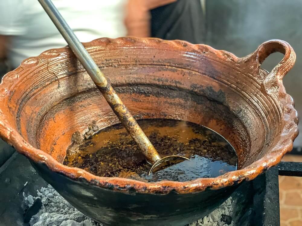 A large clay pot with braided edges and a small handle on top holds a spoon and a brown soupy mixture.