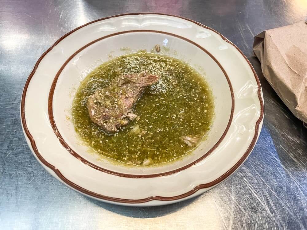 A piece of meat in a light green sauce sits on a white plate with brown design.