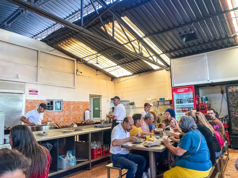 Groups of people sit at long tables in a large space under a corrugated metal roof.