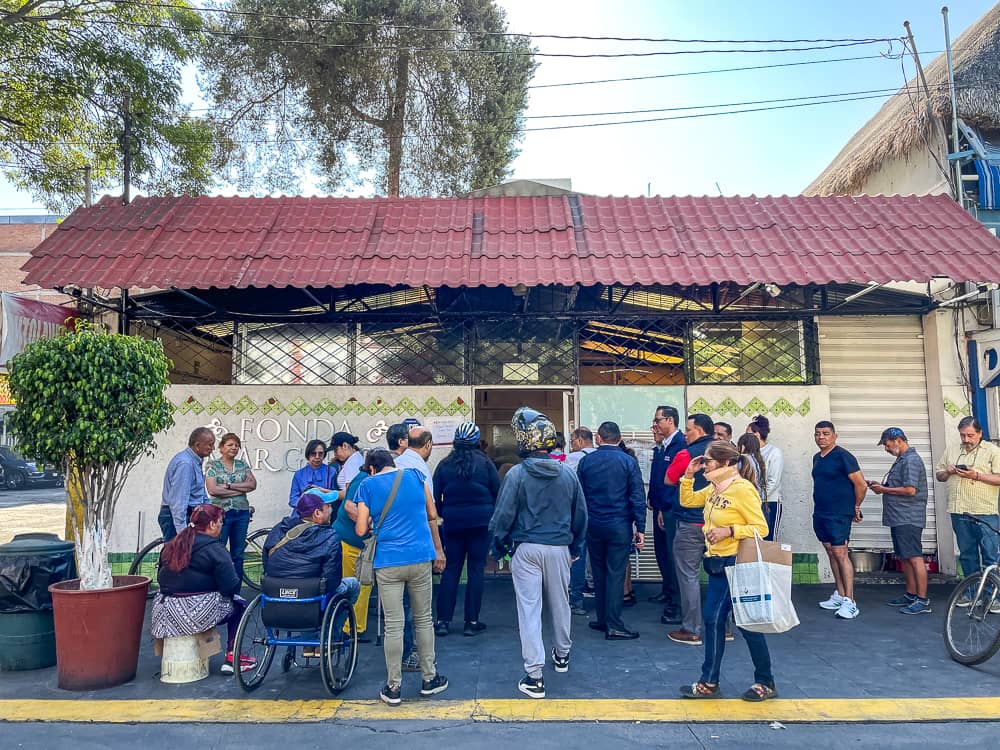 A large group of people stands in front of a small shed-like building with a red corrugated metal roof.