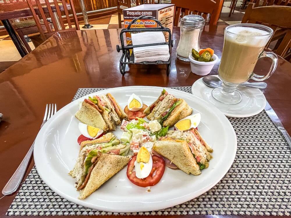 A club sandwich in quarters and tomato and hard-boiled egg garnish sits on a plate. Next to the plate there is a glass mug of coffee with foam, a napkin dispenser, a sugar shaker, and a small white bowl with pickles.