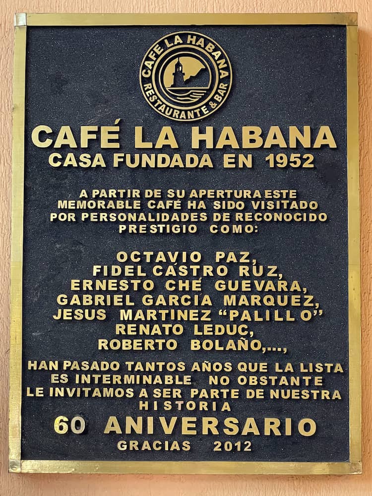 A black and gold sign that says CAFÉ LA HABANA CASA FUNDADA EN 1952 A partir de su apertura este memorable café ha sido visitado por personalidades de reconocido prestigio como: Octavio Paz, Fidel Castro Ruz, Ernesto Ché Guevara, Gabriel García Márquez, Jesús Martínez "Palillo" Renato Leduc, Roberto Bolaño Han pasado tantos años que la lista es interminable. No obstante le invitamos a ser parte de nuestra historia. 60 ANIVERSARIO Gracias 2012