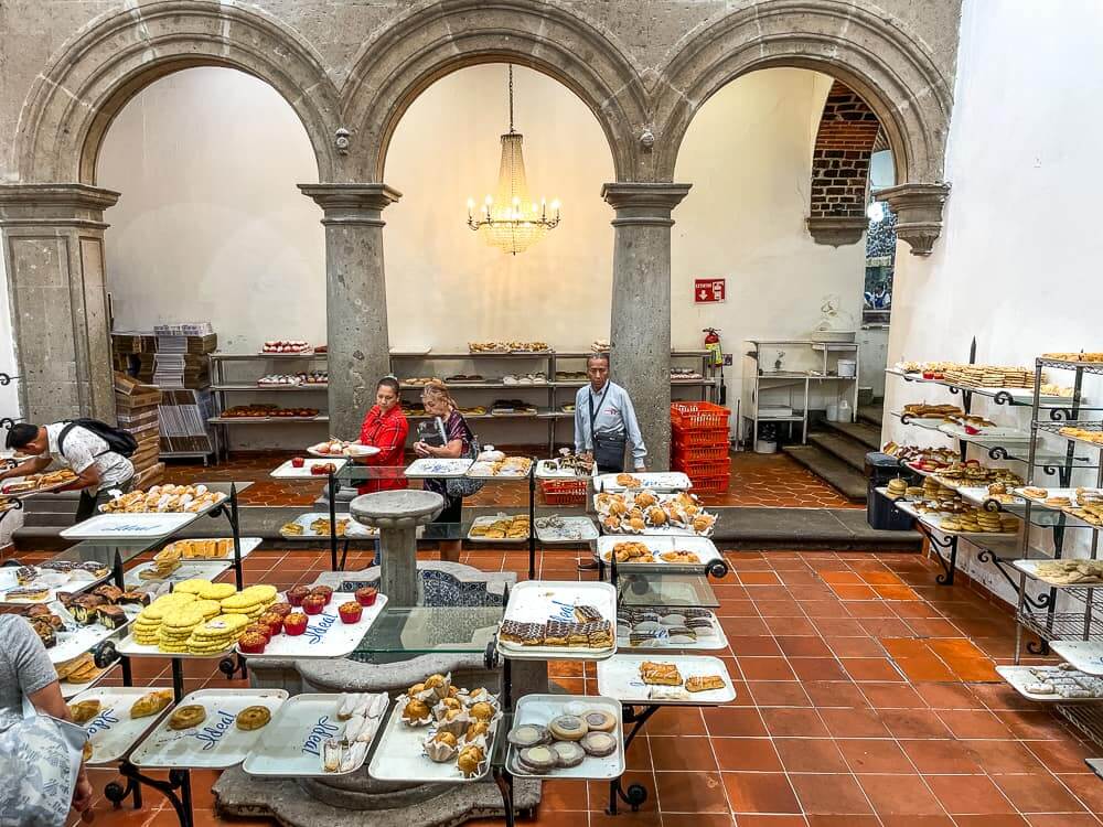 A large high-ceilinged room with three stone arches holds shelves and trays filled with pastries of all shapes and colors. People walk around looking at the pastries.