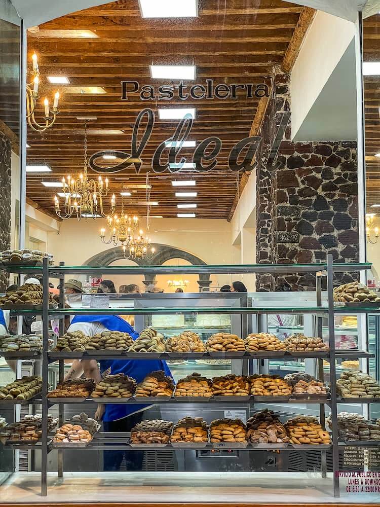 A large window looks into a well-lit shop. Stacks of pastries on shelves sit in front of the window.