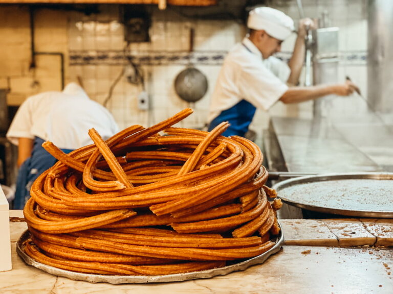 A tall circular pile of fresh churros sits on a counter. Workers dressed in white shirts and hats work the fryers.