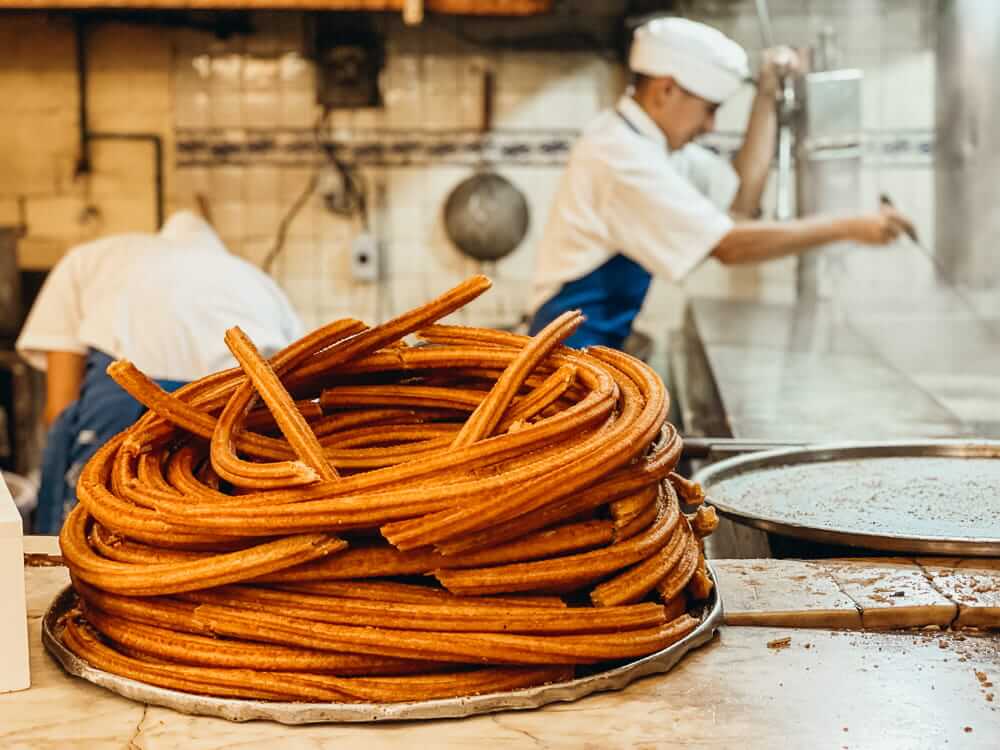 A tall circular pile of fresh churros sits on a counter. Workers dressed in white shirts and hats work the fryers.