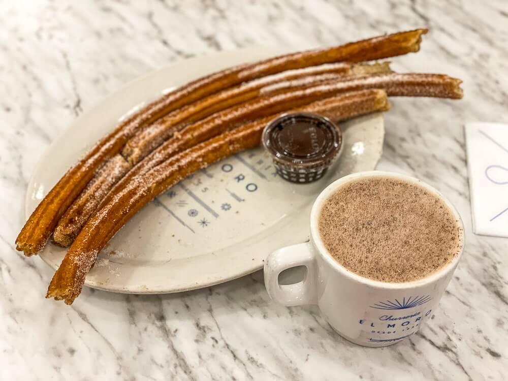 A white plate topped with four long churros and a small cup of chocolate sauce sits on a marble table. Next to the plate is a cup of foamy hot chocolate.