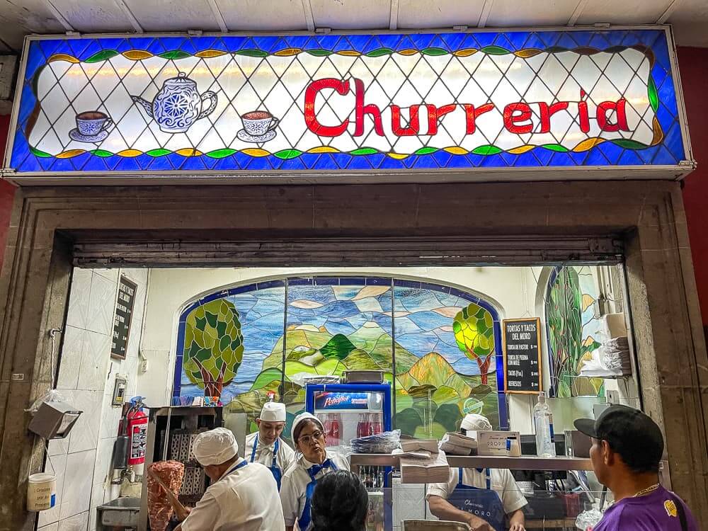 Image of a storefront bustling with workers in blue aprons and white hats. There is a blue and white stained-glass sign above with a picture of a teapot and two cups and the word Churreria in red.