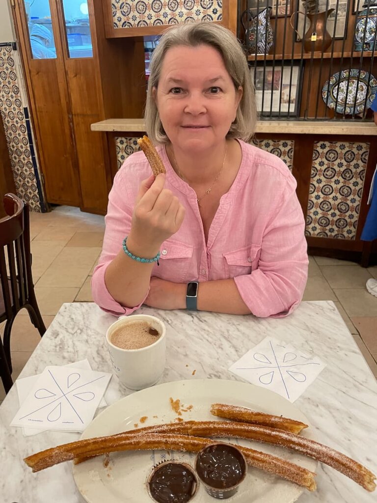 A woman in pink shirt sits at a table. She is holding a churro in her right hand. There is a plate with churros and chocolate on the table, along with a cup of hot chocolate.