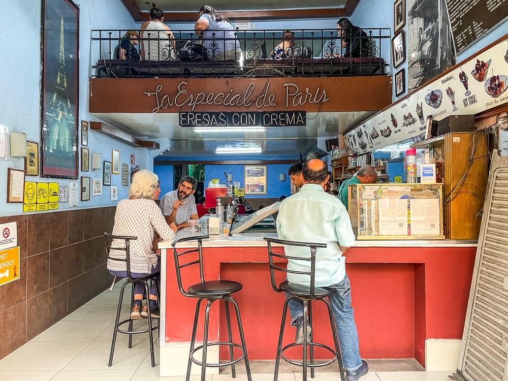 People sit on bar stools around a counter in an ice cream shop. A cursive sign that says La Especial de Paris, and below it Fresas Con Crema.