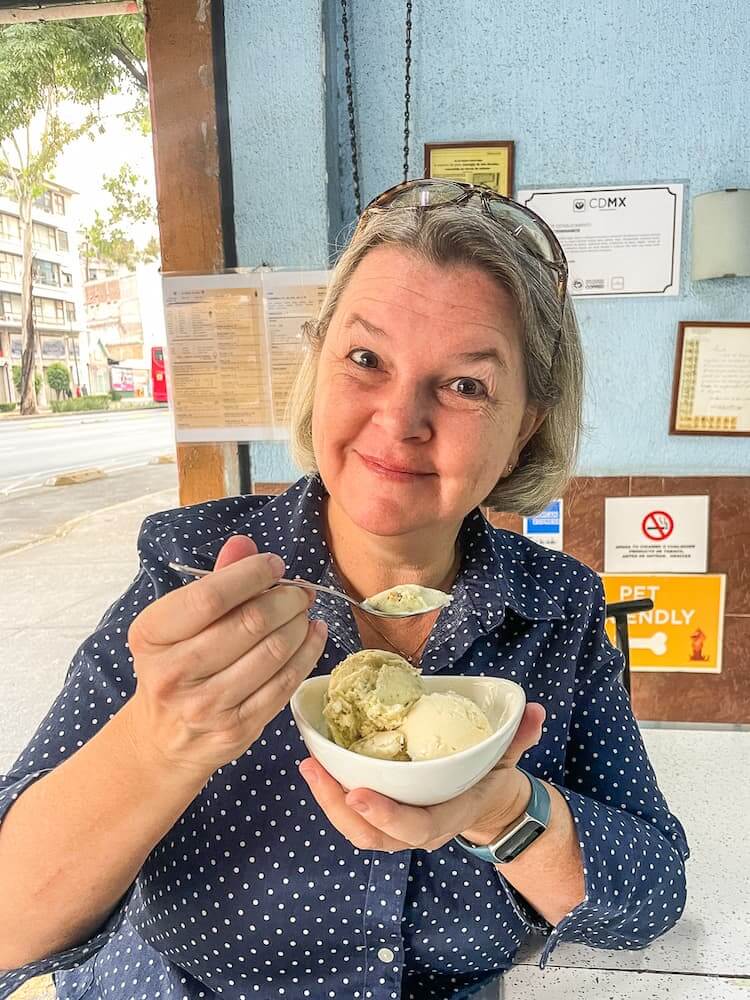 A girl in a blue shirt with small white polka dots and glasses on her head holds a spoon and a bowl with two scoops of pale ice cream.