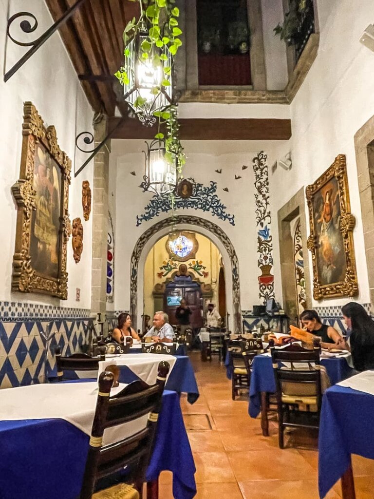 The interior of a restaurant with white walls and very high ceilings. Tables have blue and white tablecloths.