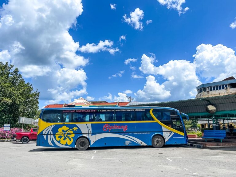 A blue bus with yellow, red and white paint is parked. The sky is blue with puffy clouds.