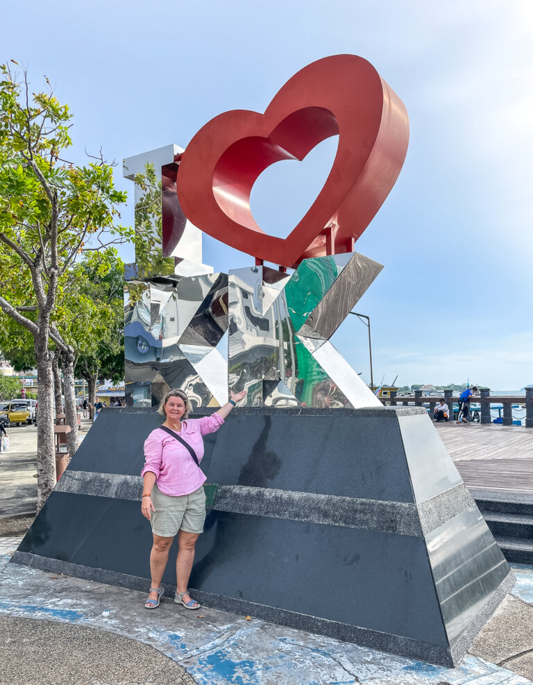 A woman in a pink shirt points her left arm at a tall metal sculpture sign that says I [HEART] KK