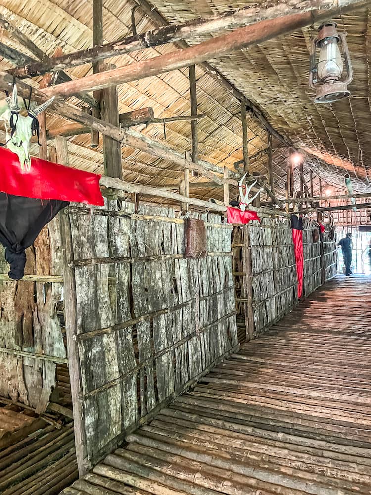 The interior of a long house made of small tree trunks. There are several rooms to the left side. 