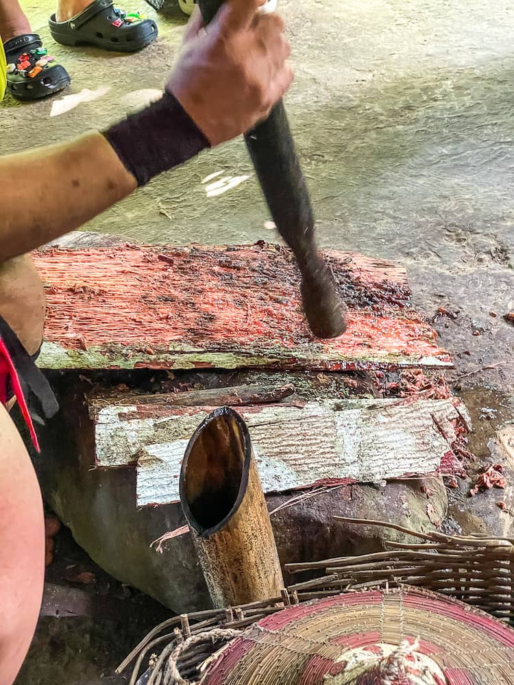 A hand holding a wooden stick pounds a red substance on a platform. 