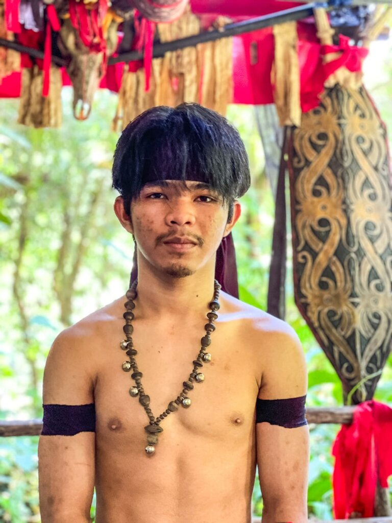 A shirtless man with black hair wearing a beaded necklace. 