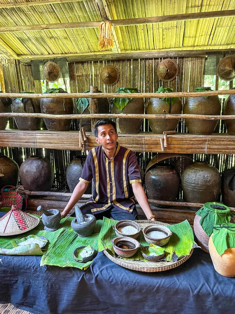 A man in a black shirt with vertical stripes sits behind a low table. On the table are various trays with bowls sitting on large green leaves.