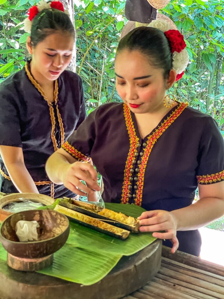 Two women wearing black shirts work on preparing food in a long bamboo vessel. 