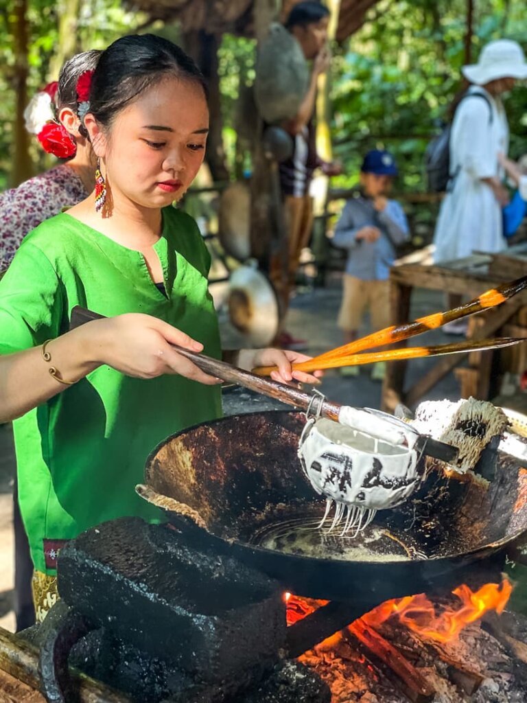 A woman wearing a bright green shirt uses a mesh scoop to drip a white batter into a pot of hot oil.