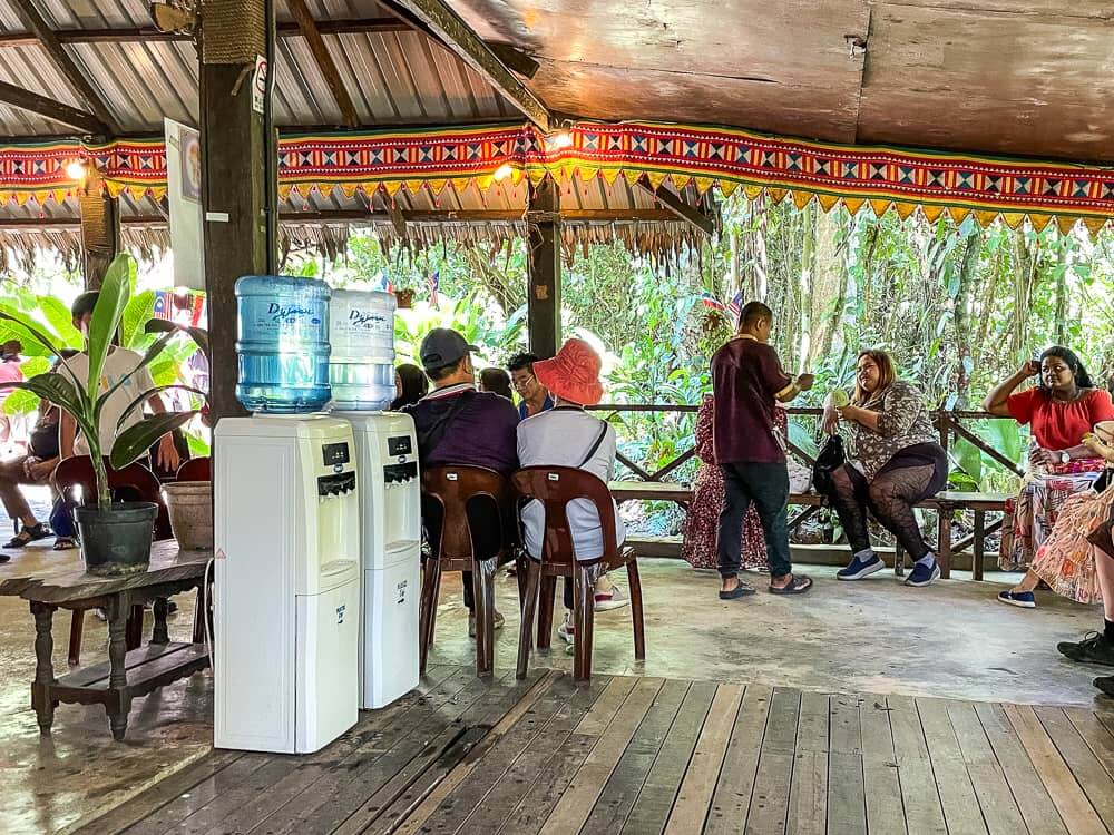 People sit on chairs on a covered wooden platform. There are two water dispensers on the left. 