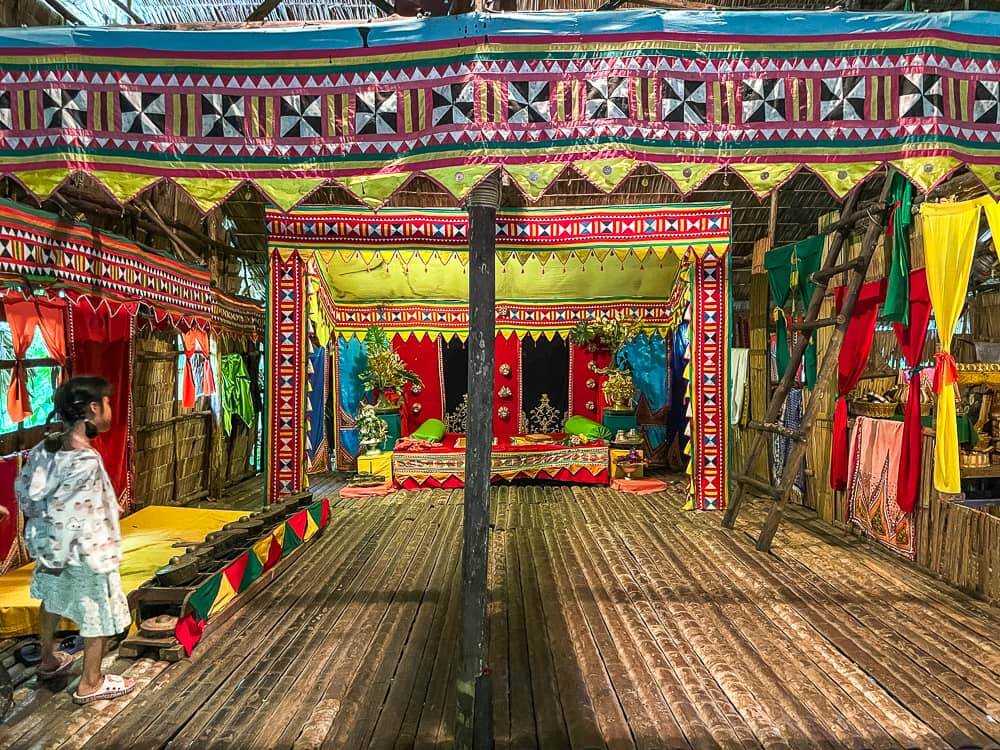 Interior view of a wooden house with colorful cloth decorations hanging from the ceiling. There is a wooden pillar in the middle of the room.