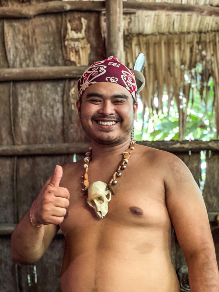 A smiling shirtless man wearing a headress and a necklace with a small animal skull holds up his right hand with a thumbs up