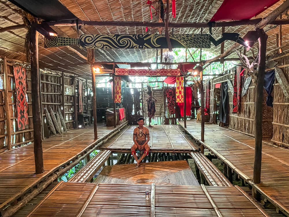 A man sits on the floor in a large wooden house. The floor in the center of the house is slightly sunken. 