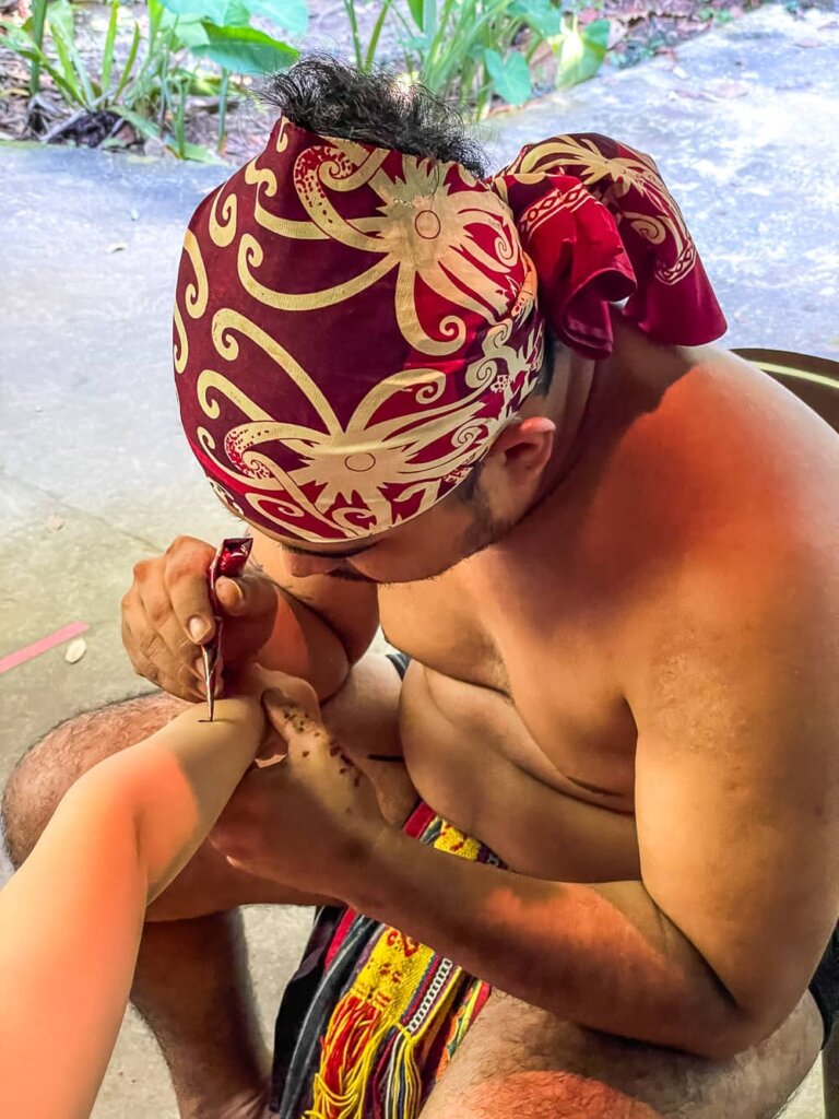 A shirtless man with a red head-wrap uses henna to draw on a small arm. 