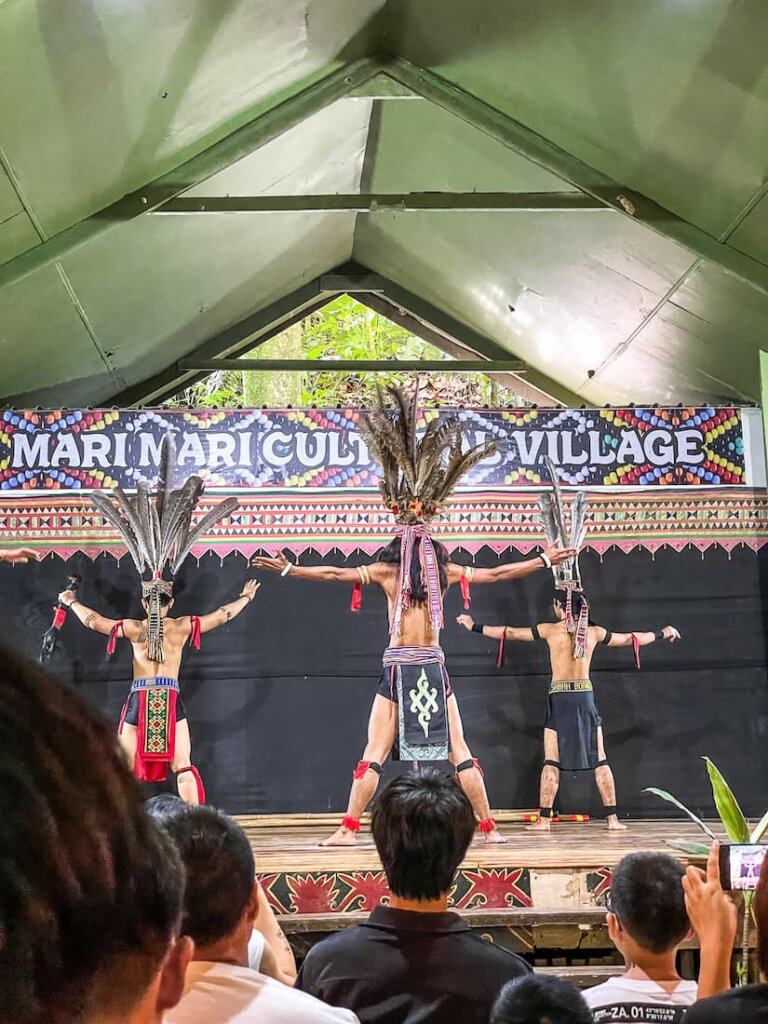 Three men in elaborate feather headdresses with their arms out stand on a stage. 