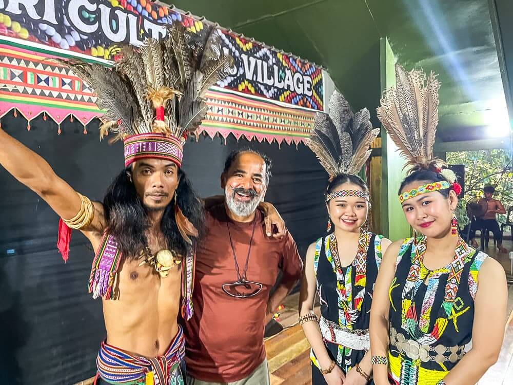 Four people pose for a picture. The man on the left and the two women have an elaborate feathered headdress. 